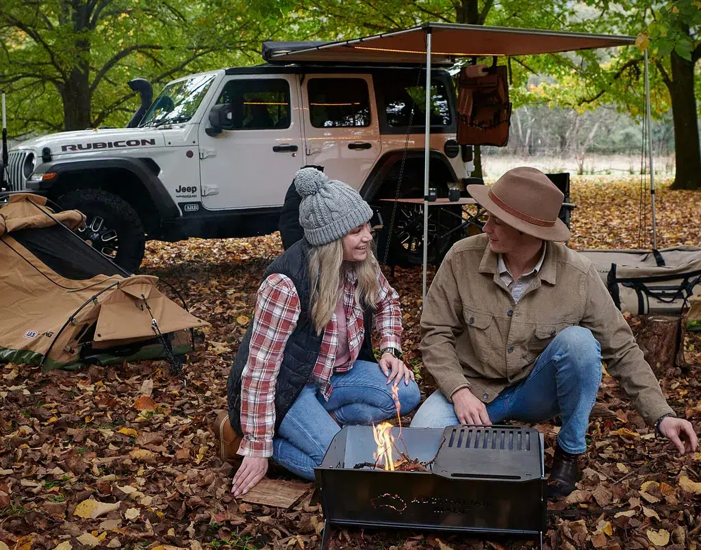 Couple camping in a fall forest sits by a small campfire with a white Jeep Wrangler Rubicon and tents nearby? under an awning.